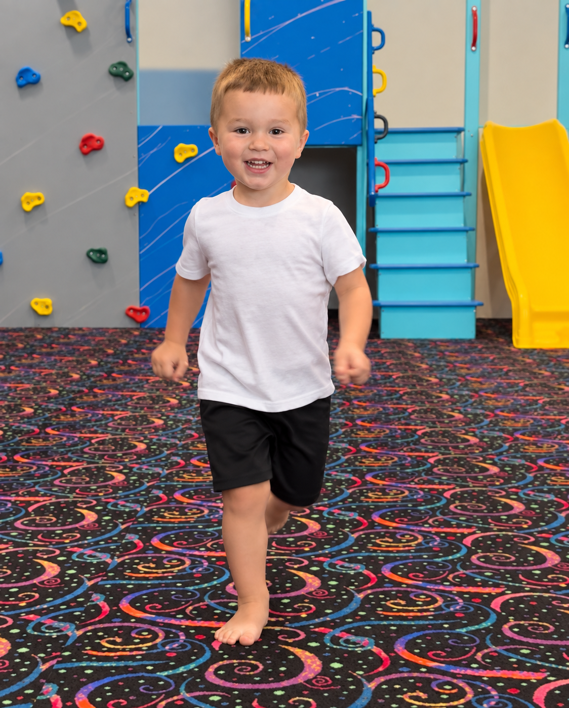 A smiling child running through The Grove play gym beside the climbing wall and yellow slide